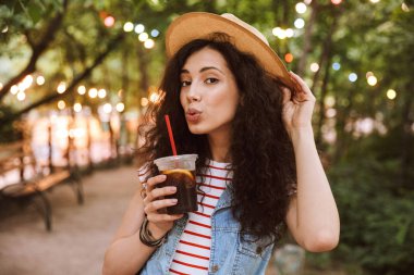 Esmer genç kadın 18-20 giyen yaz hasır şapka güzel aydınlatmalar ile parkın içinden yürürken plastik bardak soğuk çay içme portresi