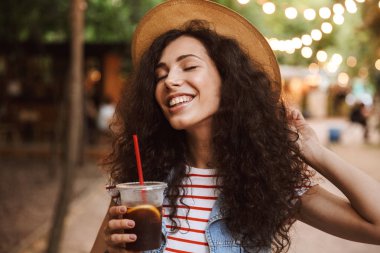 Fotoğraf Renkli lambalar ile parkta yürürken plastik bardak soğuk çay içme mutlu sevimli kadın 18-20 giyen yaz hasır şapka
