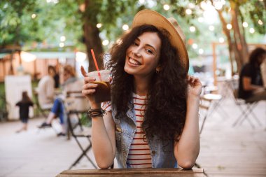 Görüntü güzel mutlu kadın 18-20 giyen yaz hasır şapka park kafede istirahat ederken plastik bardak soğuk çay içme
