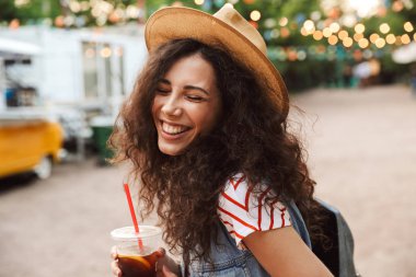 Neşeli sevimli kadın 18-20 diğer açık için şirin yer ile yürürken plastik bardak soğuk çay içme yaz hasır şapka kıvırcık kumral fotoğrafı