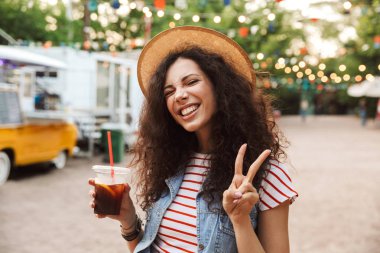 Görüntü mutlu güzel kadının 18-20 yaz hasır şapka gülümseyen ve plastik kap. barış işareti gösterilen süre açık soğuk çay içme kıvırcık kumral