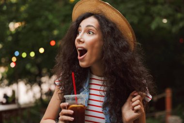 Mutlu kadın 18-20 hasır şapka bir kenara seyir açık ağız ile yeşil park içinde kalan sırasında plastik bardak içecek yudumlarken giyen portre closeup heyecanlı