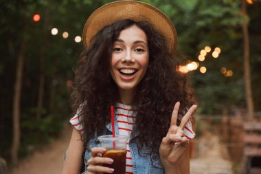 Portre tatlı esmer güzel kadın 18-20 plastik bardak soğuk çay içme ve dinlenme yeşil park sırasında barış işareti gösteren hasır şapka