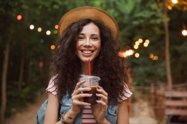 Plastik bardak içecek içme ve renkli lambalar arka plan ile yeşil parkta yürüyüş yaparken bakıyorsun görüntü saman neşeli yaz kadın 18-20 giyen şapka
