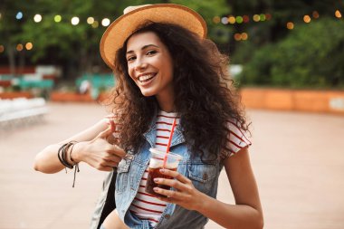 Şık beyaz kadın 18-20 renkli lambalar arka plan ile hipster spot ya da modern Park'ta dinlenme sırasında plastik bardak içecek içme kıvırcık kumral fotoğrafı
