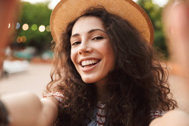 Genç esmer kadın 18-20 gülümseyen ve yenilikçi yer veya renkli aydınlatma arka plan ile modern park yürürken selfie fotoğrafta kıvırcık kumral