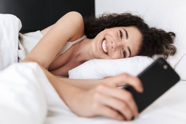 Mutlu kafkas kadın 20'li siyah kıvırcık saçlı otel odasında beyaz yastık üzerinde yatakta yalan ve gülümseme ile cep telefonu görünümlü görüntü