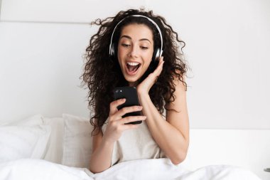 Neşeli genç kadın 20'li yaşlarda portresi akıllı telefon ve kulaklık Beyaz yatakta evde müzik dinlerken şarkı