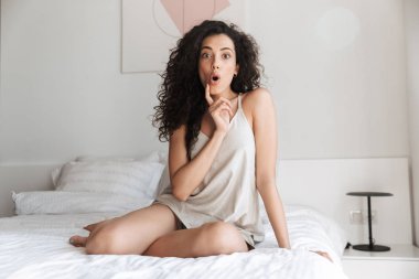 Sabah yatak beyaz keten evde oturan ve açık ağzı ile sürpriz ifade heyecanlı kıvırcık kadın görüntüsünü