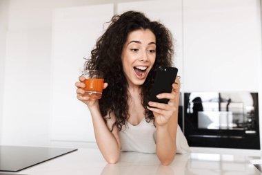 Hayret Avrupa kadın 20'li yaşlarda kıvırcık saçlı mutfak ve siyah cep telefonu kullanıyorum ve heyecan ifade suyu içme ipek eğlence giyim fotoğrafı