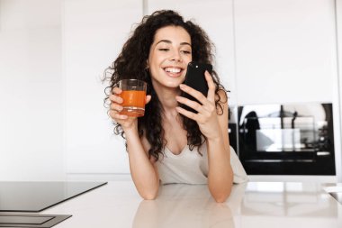 Güzel taze kadın 20'li yaşlarda kıvırcık saçlı mutfakta suyu içme ve gülümseme ile siyah cep telefonu kullanarak ipek eğlence giyim fotoğrafı