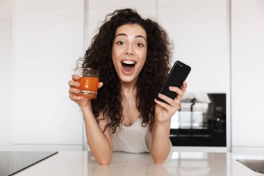 Mutfakta suyu içme ve siyah smartphone tutarak açık ağız ile kameraya bakarak Avrupa kıvırcık kadın 20'li yaşlarda giyen ipek eğlence giyim görüntü