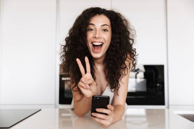 Güzel mutlu kadın 20'li yaşlarda mükemmel ile gülümseyen ve siyah smartphone tutarken barış işaret parmaklarıyla gösteren ipek eğlence giyim kıvırcık kumral