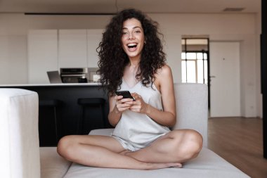 Kıvırcık saçlı modern Flat kanepede otururken cep telefonu kullanarak ipek eğlence giyim heyecanlı genç kadın fotoğrafı
