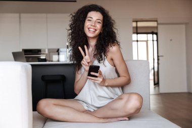Dairede oturan ve süre kanepede oturan ve barış işareti gösterilen cep telefonu kullanarak sevimli gülümseyen kadın fotoğrafı