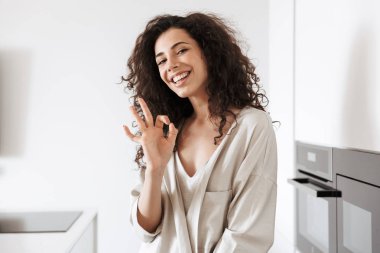 Uzun siyah saçlı kameraya gülümseyen ve evde mutfakta dururken ok işaret gösteren ipek eğlence giyim mutlu genç kıvırcık kadın fotoğrafı