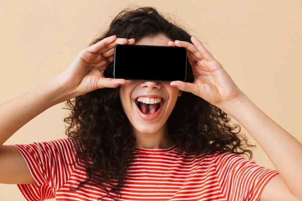Gözleri kapsayan cep telefonuyla görüntü gösterilen bej renkli arka plan üzerinde izole duran sevimli genç kıvırcık kadın görüntüsünü.