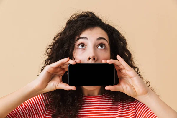 Ekran cep telefonu tarafından ağız kapsayan gösterilen bej renkli arka plan üzerinde izole duran şok genç kıvırcık kadın görüntüsü. Bir kenara arıyorsunuz.