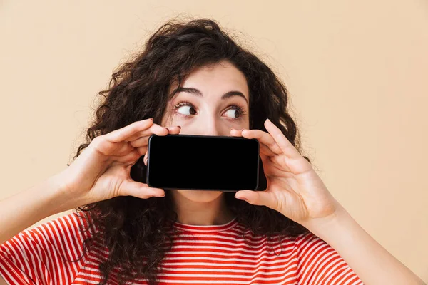Ekran cep telefonu tarafından ağız kapsayan gösterilen bej renkli arka plan üzerinde izole duran şok genç kıvırcık kadın görüntüsü. Bir kenara arıyorsunuz.