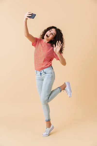 满长的肖像一个快乐的年轻女孩采取自拍手机 而跳跃和挥舞着在米色背景下孤立 — 图库照片
