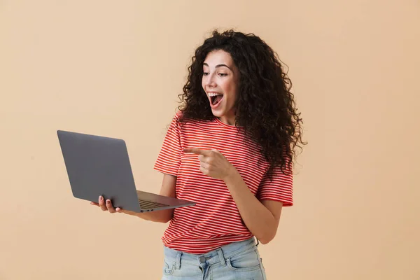 形象的兴奋漂亮的年轻卷曲的女人在米色背景下使用膝上型电脑指向 — 图库照片