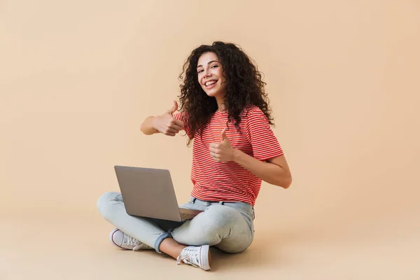 图像的快乐漂亮的年轻卷曲的女人 在米色背景下使用笔记本电脑 看相机显示竖起大拇指 — 图库照片