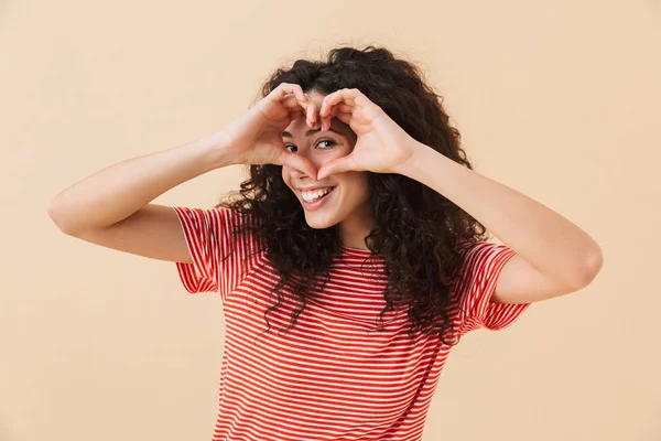 Kamera seyir bej duvar arka plan üzerinde izole mutlu genç kadının fotoğrafını jest seviyorum kalp yapmak.