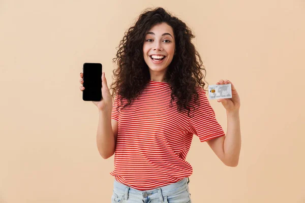 Sevimli Genç ve güzel kıvırcık kadın cep telefonu kredi kartı tutan görüntüsünü gösterilen bej renkli arka plan üzerinde izole görüntüsünü.