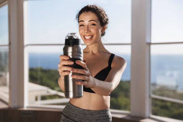 Cheerful Young Sportswoman Showing Water Bottle While Standing Window Indoors — Stock Photo, Image
