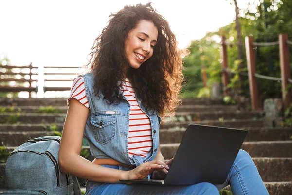 欧洲黑发妇女的肖像 微笑着坐在楼梯上的夏日公园和使用银笔记本电脑 — 图库照片