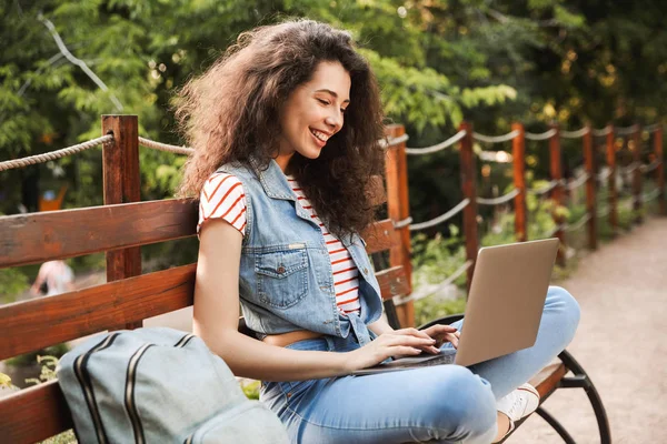 Güneşli günde yeşil parkta bankta oturmuş ve istimal gümüş laptop sırt çantası olan esmer kadın görüntüsünü