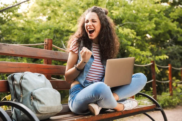 照片的激动兴奋的女人20S 卷曲的头发紧握拳头 而坐在绿色公园的长凳上 学习或工作的银色笔记本电脑 — 图库照片