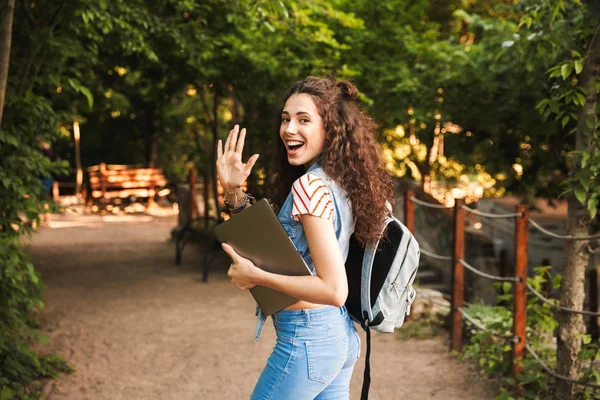 Dostu çekici kadın portresi 18-20 giymiş sırt çantası kamera sallanan el elinde gümüş dizüstü bilgisayar ile yeşil parkın içinden yürürken