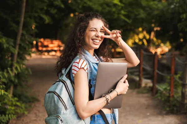 Dizüstü bilgisayar açık havada Park'ta yürüyordunuz barış hareketi gösterilen tutan mutlu genç güzel kadın görüntüsü. Görünümlü kamera.