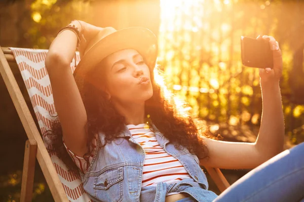 Açık havada park yapmak selfie öpücük üfleme cep telefonuyla konuşan genç güzel bir kıvırcık kadın görüntüsünü.