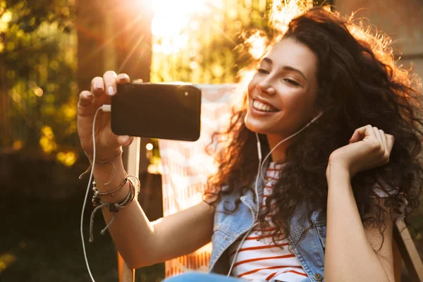 Selfie tarafından cep telefonu kulaklık ile müzik yapmak imge-in şirin genç güzel kıvırcık kadın açık havada park.