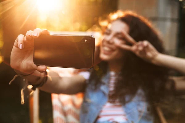 屋外公園でかわいい若い美しい巻き毛女性像は平和のジェスチャーと携帯電話で Selfie を作る — ストック写真