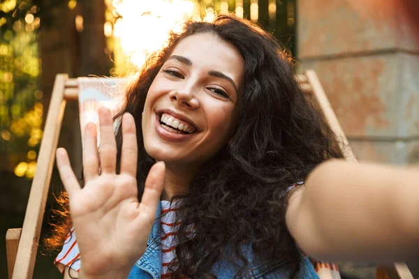 18-20 kameraya bakarak ve lounge sandalye açık ve çekici selfie fotoğraf otururken el sallayarak gülümseyen kıvırcık kız memnun
