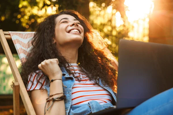 Photo of joyful brunette woman 18-20 clenching fist in joy while sitting in lounge chair during rest in park on sunny day and using silver laptop with surprise