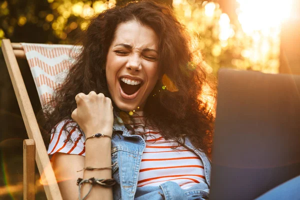 Photo of ecstatic brunette woman 18-20 clenching fist and screaming while sitting in deck chair during rest in park on sunny day and using silver laptop