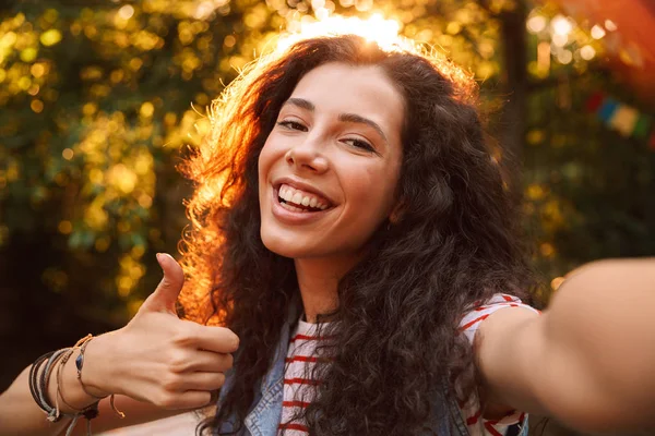 Memnun gülümseyen kıvırcık kız 18-20 kameraya bakarak ve açık yürüyüş ve alma selfie başparmak gösterilmesini ağaçlar arka plan ile fotoğraf