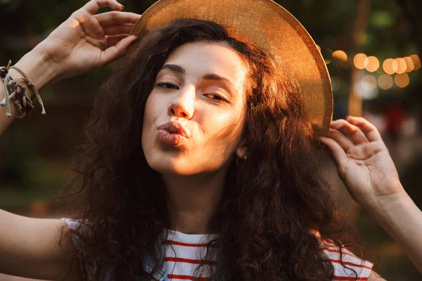 Esmer çok mutlu kadın 18-20 giyen yaz hasır şapka ağaçlar arka plan ile güneşli gün parkta yürürken hava öpücük sana üfleme closeup fotoğraf