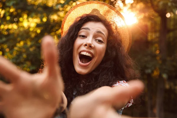 Fotoğraf gülümsemek gülümseyen ve ağaçlar arka plan ile güneşli gün parkta yürürken elinde kamera için yayılan genç kadın 18-20 giyen yaz hasır şapka