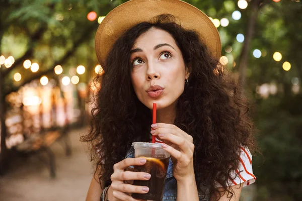 Esmer güzel kadın 18-20 giyen yaz hasır şapka aydınlatma ile parkın içinden yürürken plastik bardak soğuk çay içme closeup fotoğraf