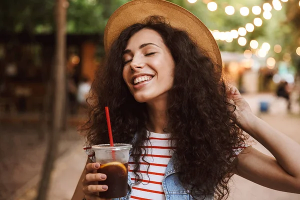 Fotoğraf Renkli lambalar ile parkta yürürken plastik bardak soğuk çay içme mutlu sevimli kadın 18-20 giyen yaz hasır şapka