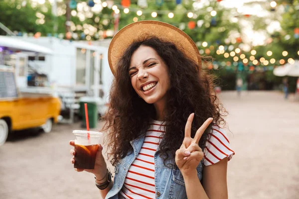 Görüntü mutlu güzel kadının 18-20 yaz hasır şapka gülümseyen ve plastik kap. barış işareti gösterilen süre açık soğuk çay içme kıvırcık kumral