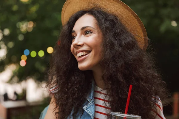 Photo of happy european woman 18-20 wearing straw hat laughing and looking aside while drinking beverage from plastic cup during rest in green park