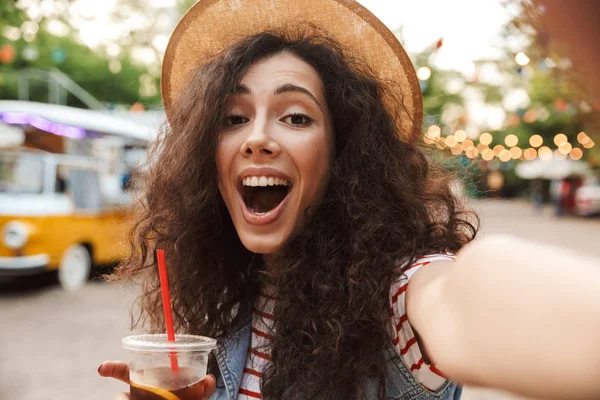 笑顔の Selfie 写真を取っている間屋外のプラスチック カップから冷たいお茶を飲む夏麦わら帽子を身に着けている黒い巻き毛の興奮若い女性 のイメージ — ストック写真