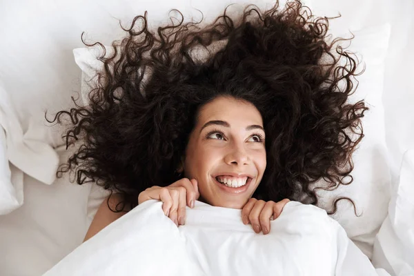 Yukarıdan portresi siyah kıvırcık saçlı yukarı mutlu gülümseme ile beyaz keten yatakta yatan süre seyir ile neşeli kadın 20'li
