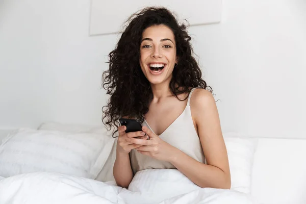 Portrait of happy woman sitting in bed after sleep on white clean linen at bedroom and smiling while holding mobile phone